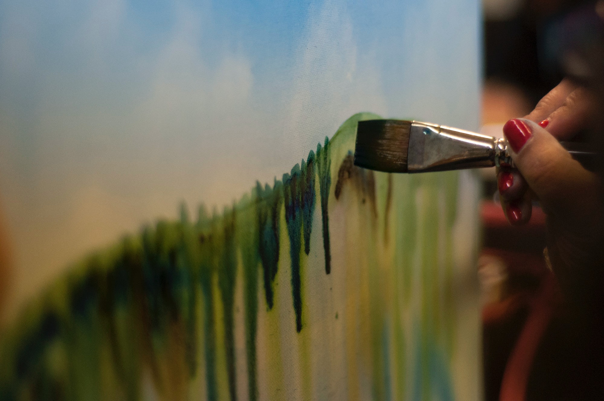 picture shows a paintbrush being used to paint on a canvas showing a blue sky with clouds and green paint showing a landscape 